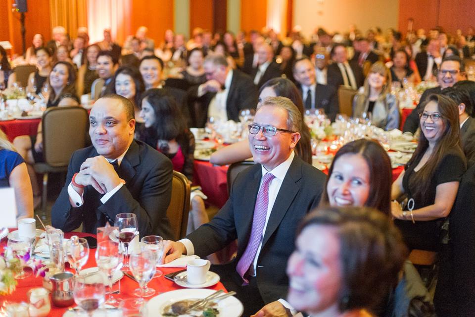 Gala dinner audience at round tables with red tablecloths, smiling attendees in formal attire in a hotel ballroom