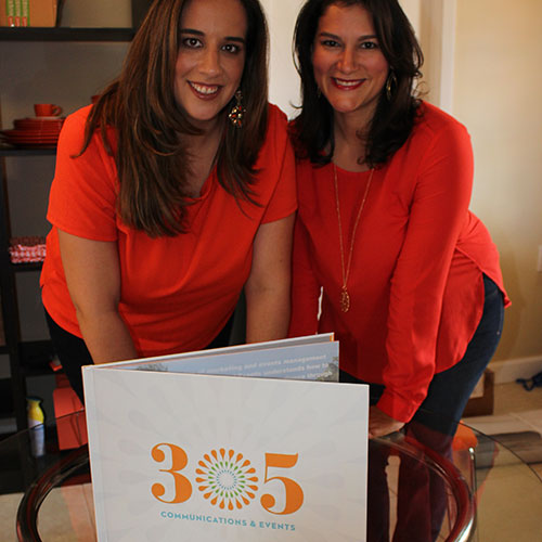 Two women in matching orange tops holding a 305 Communications & Events branded portfolio