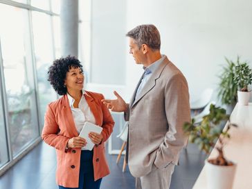 Woman in orange blazer with tablet speaking with an older colleague in a bright office hallway