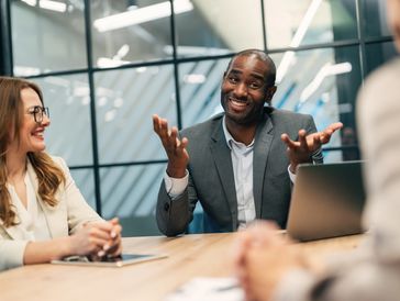 A man in a suit gesturing expressively while smiling in a meeting with colleagues