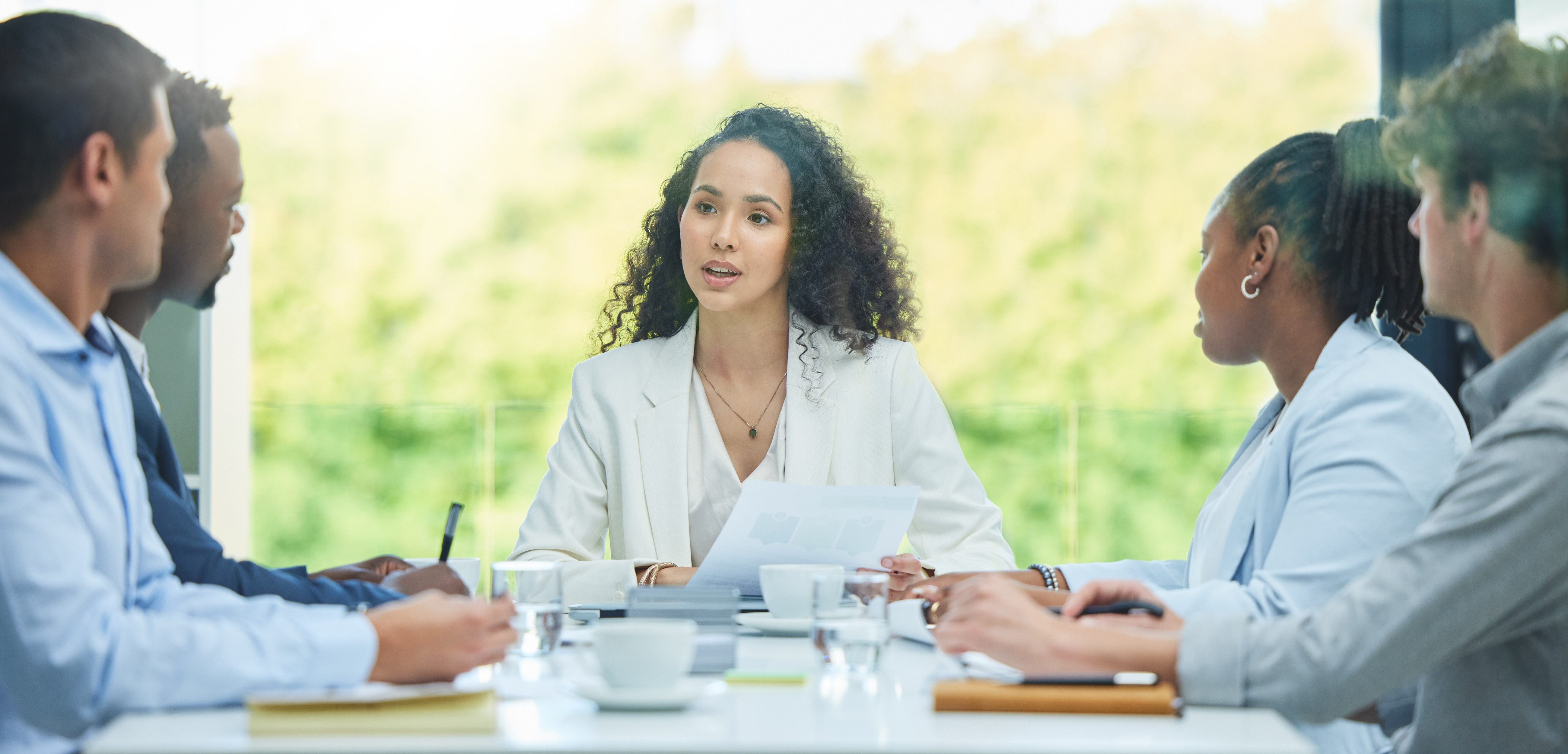 Woman in a white blazer leading a discussion with colleagues around a bright conference table