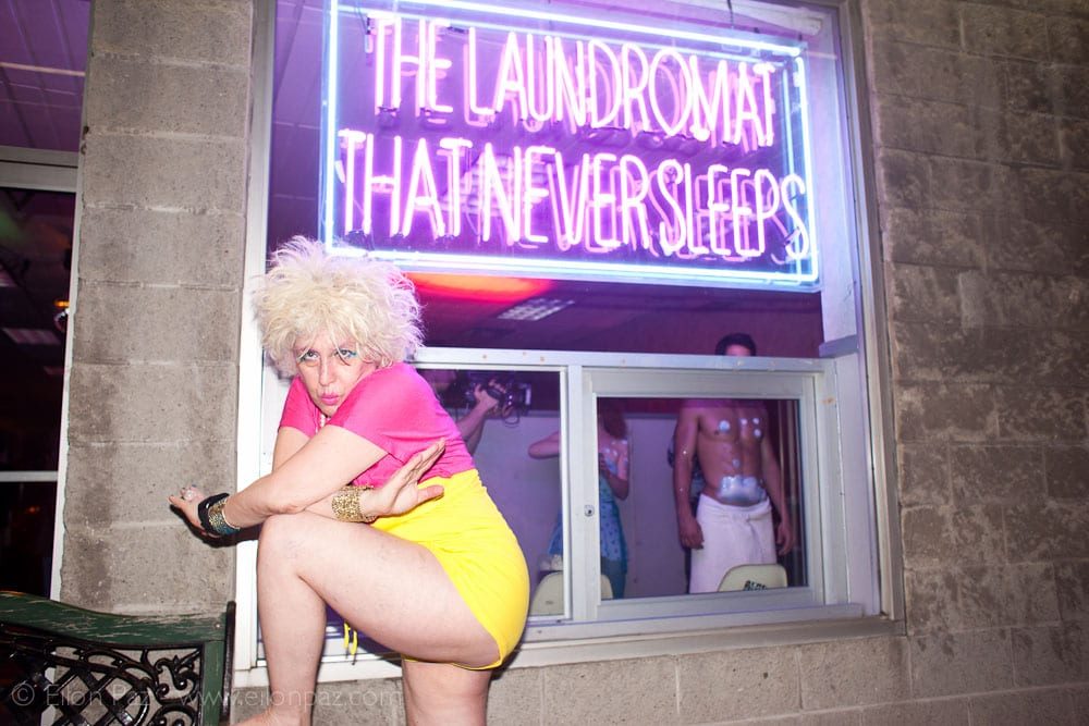 Performer beneath a neon 'The Laundromat That Never Sleeps' sign