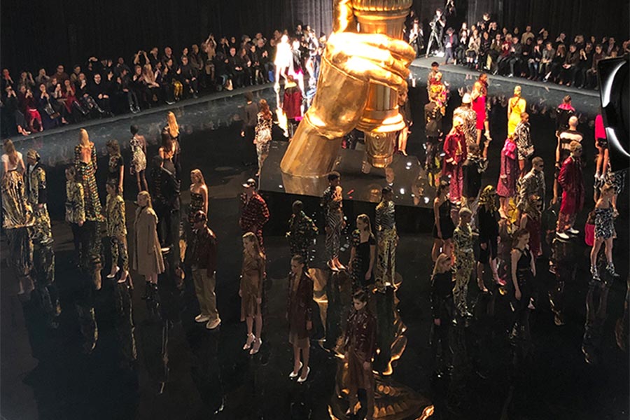 Fashion runway show finale with models around a gold sculptural centerpiece