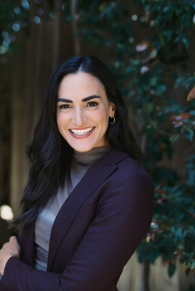 Alexandra Modon, smiling with arms crossed, outdoor foliage behind her