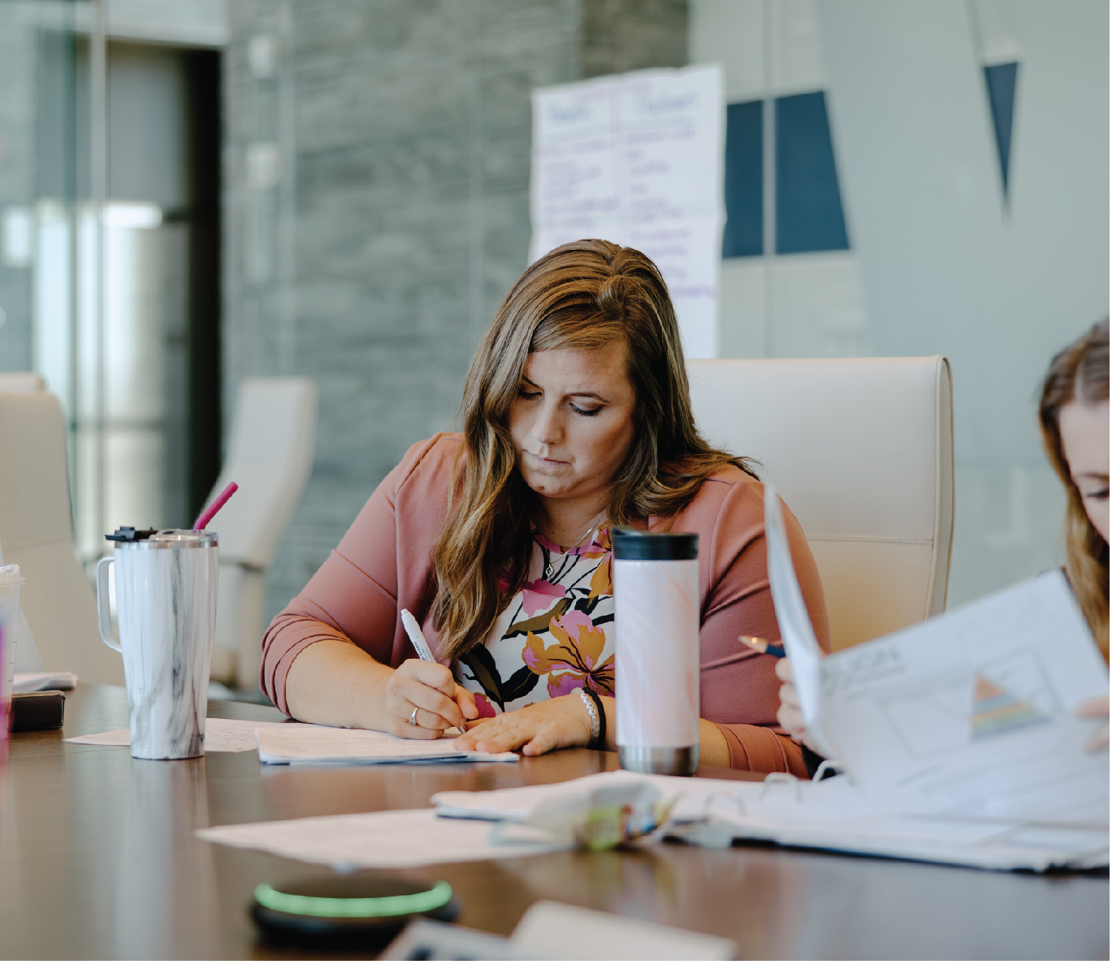 Woman in a pink blazer taking notes at a conference table while a colleague reviews papers