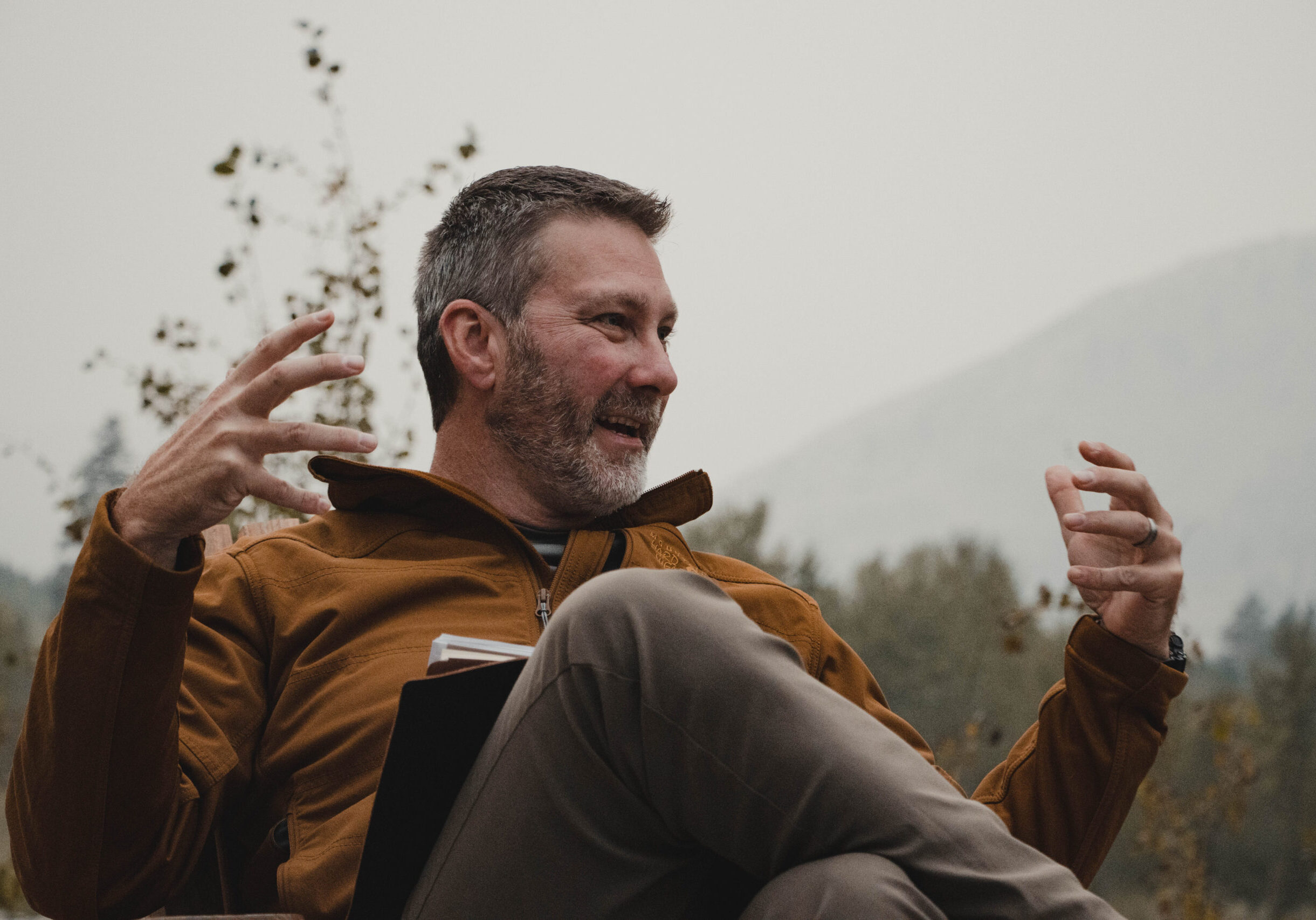 Eric Camfield, seated outdoors gesturing with both hands, mountains behind