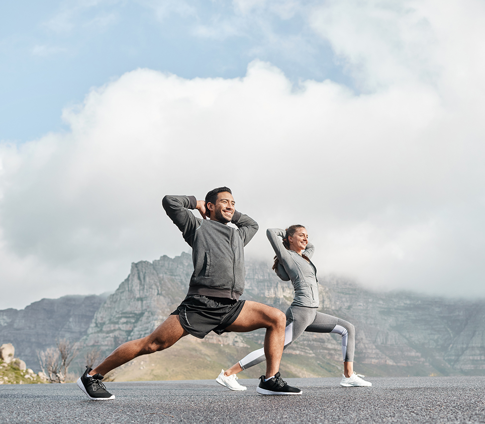 A man and woman doing lunge stretches outdoors with a mountainous backdrop