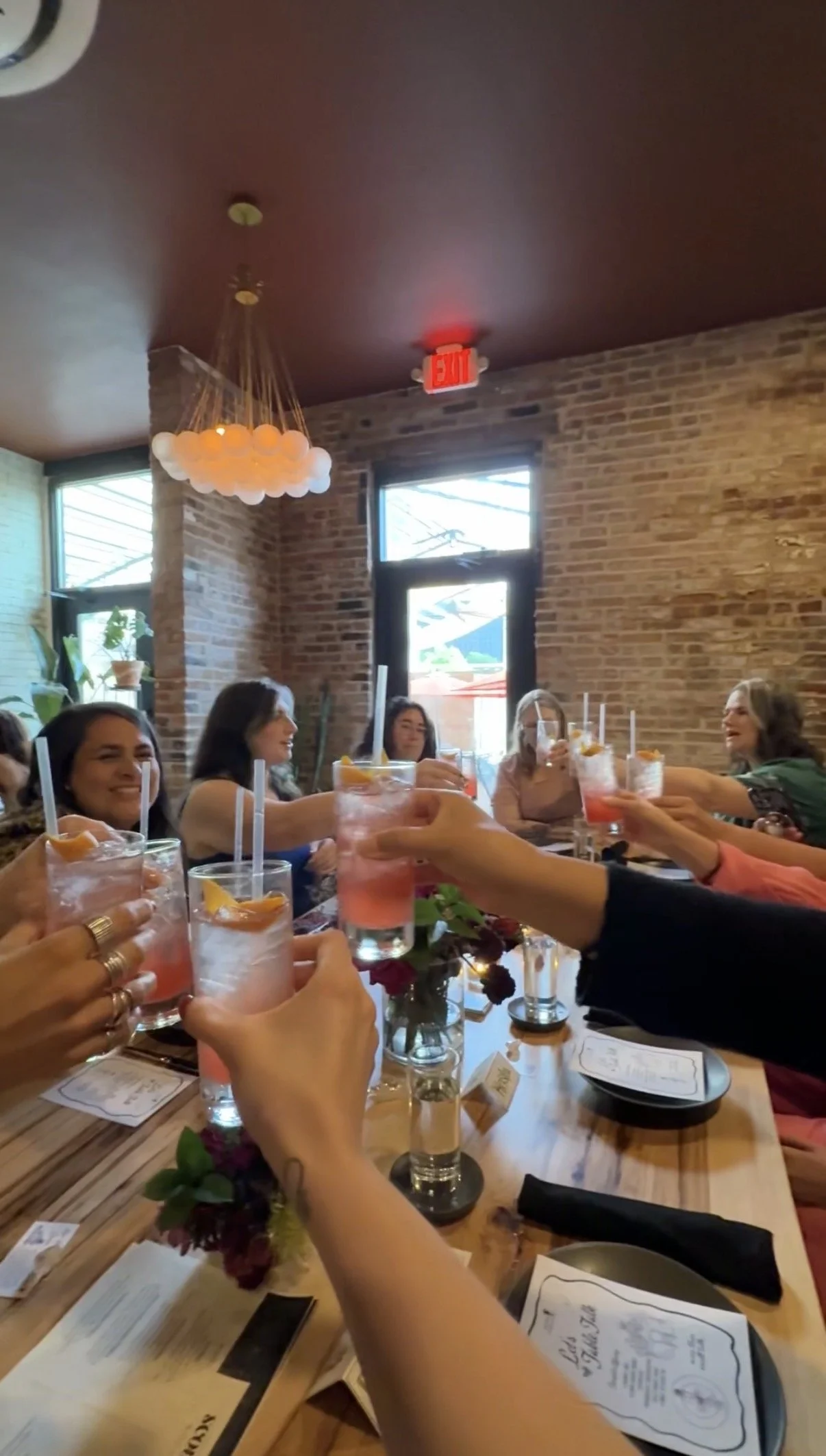 Women toasting pink cocktails at a long wooden table in a brick-walled restaurant