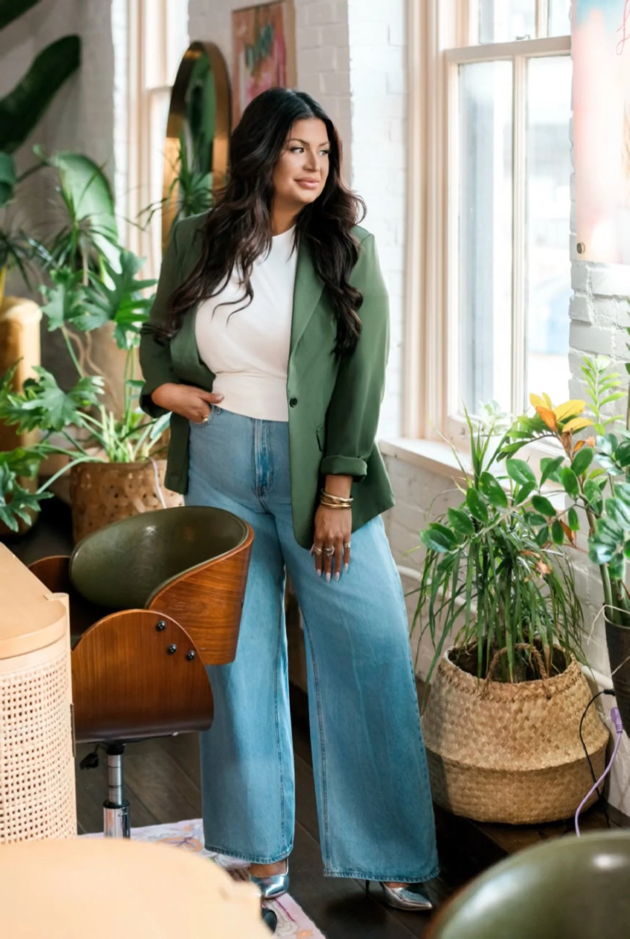Angela Berra in a green blazer standing by a window in a plant-filled interior