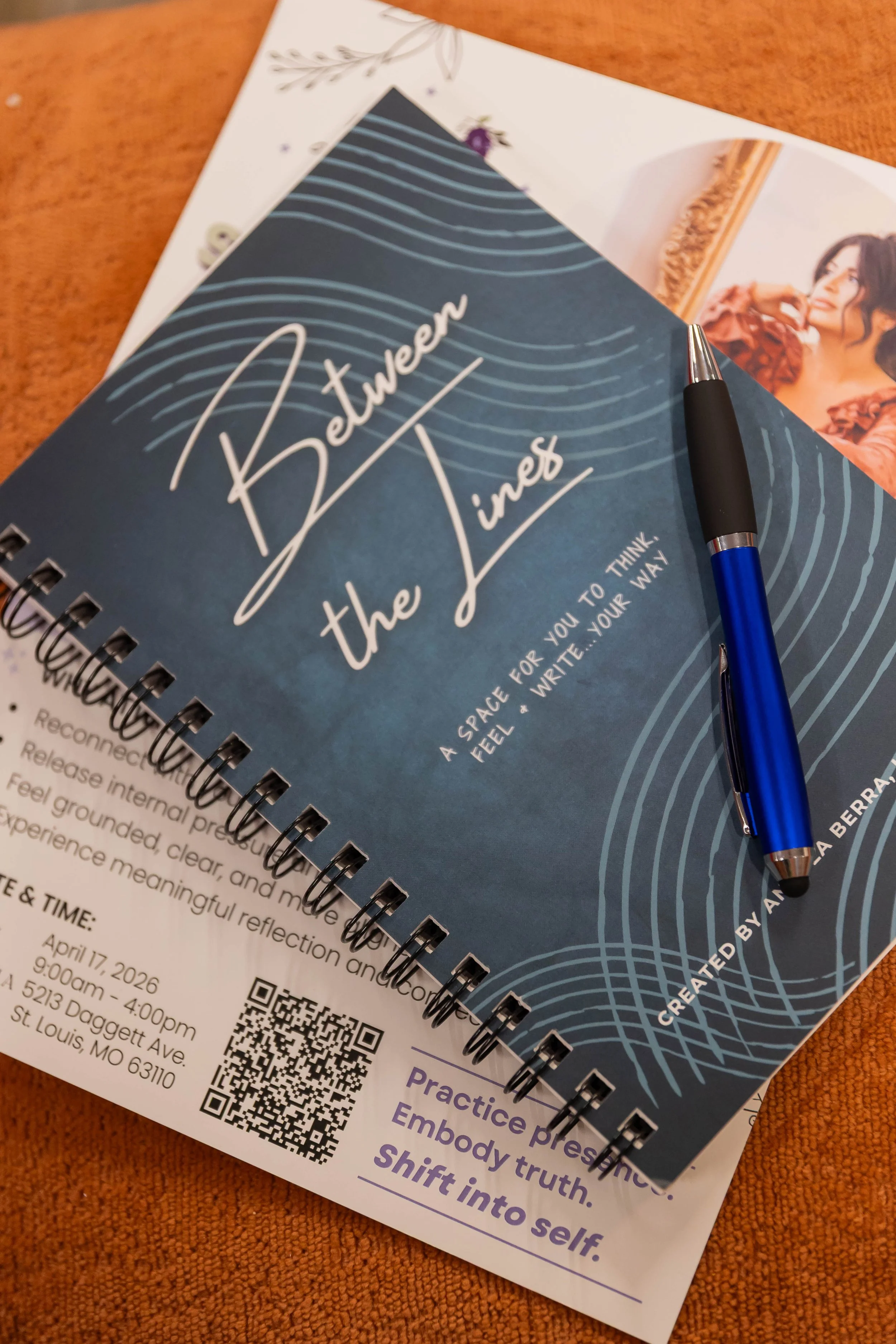 The Between the Lines spiral journal with a blue pen resting on an event flyer