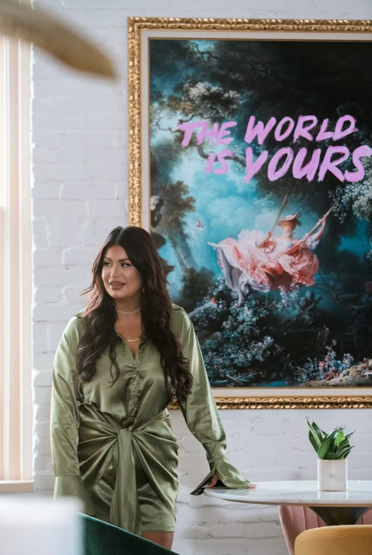 Angela Berra in a green satin dress beside a framed art print reading The World Is Yours