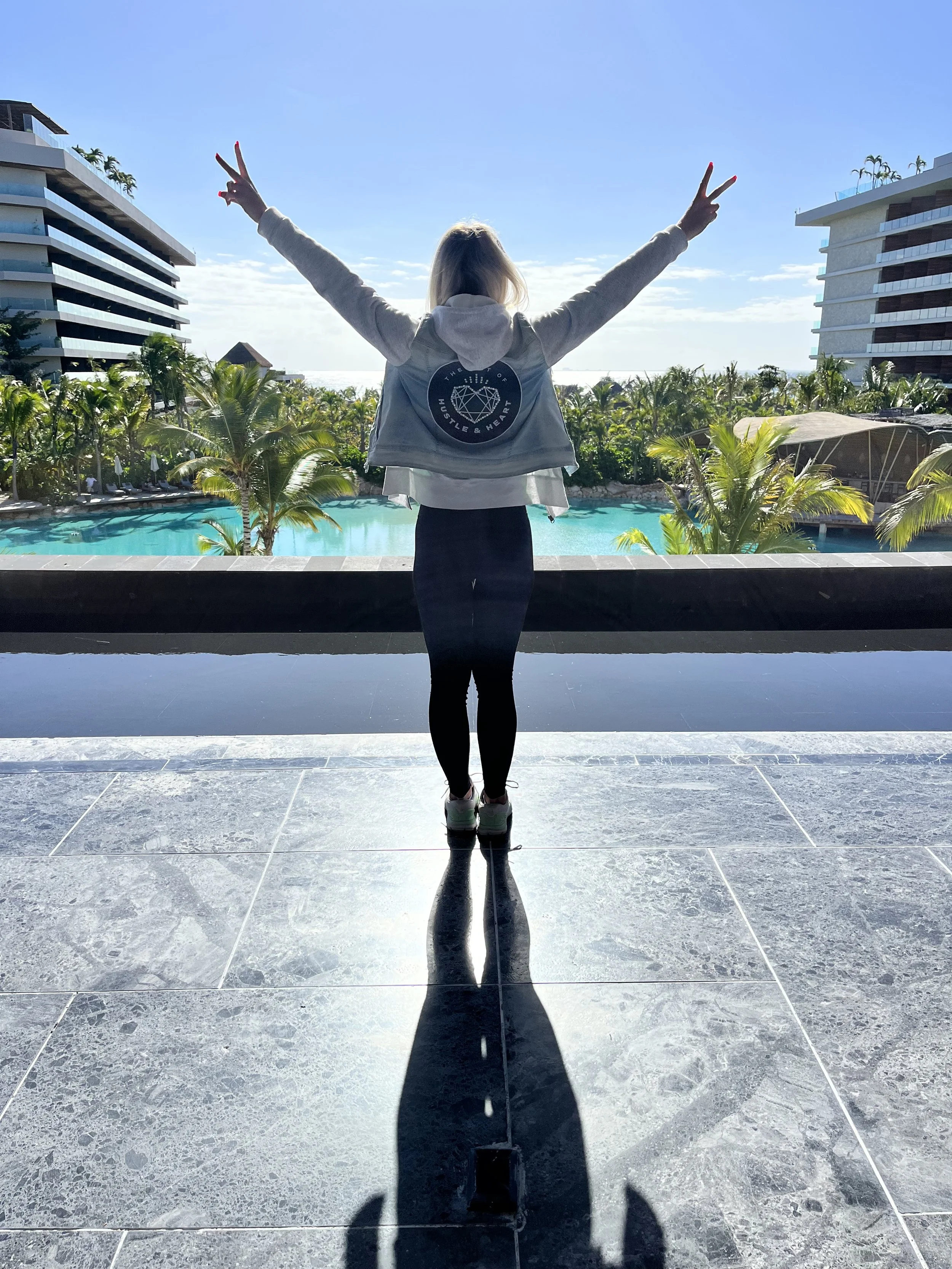 Person wearing a Hustle & Heart branded hoodie, arms raised in celebration facing a resort pool