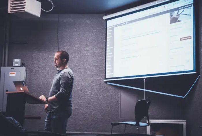 Presenter in a grey sweater at a projector screen in a dark conference room