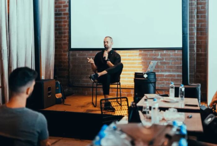 Presenter speaking on a small stage with a microphone to an audience in a brick-walled venue