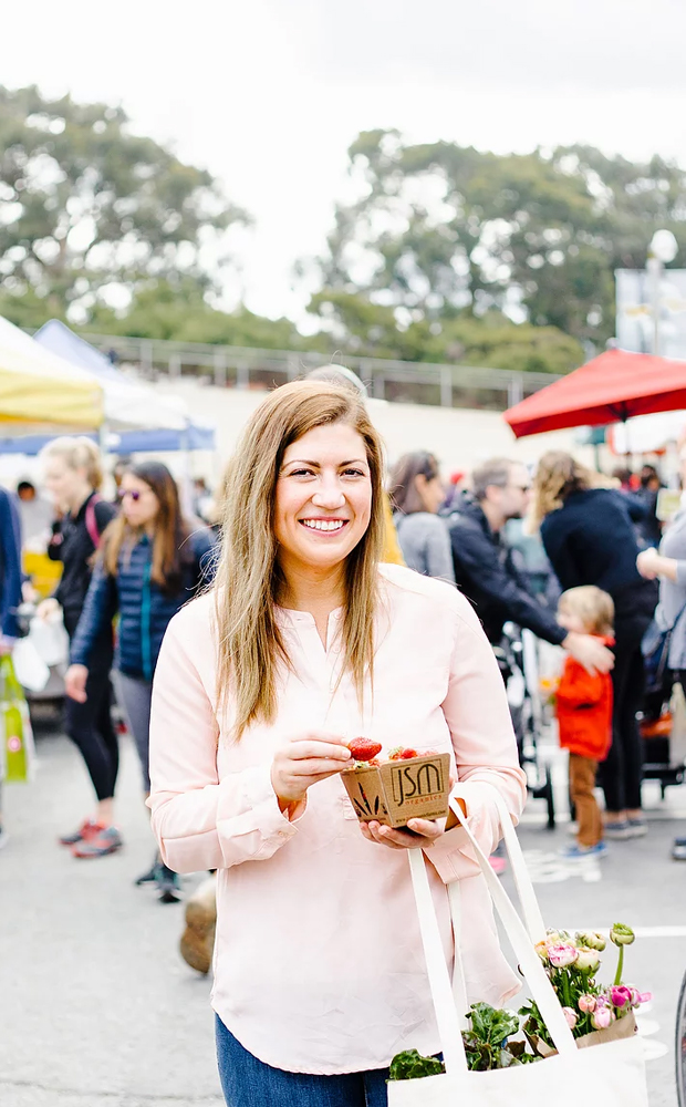 Hayley, founder of A Wholistic Life, in a pink blouse holding a box of strawberries at a busy farmers market
