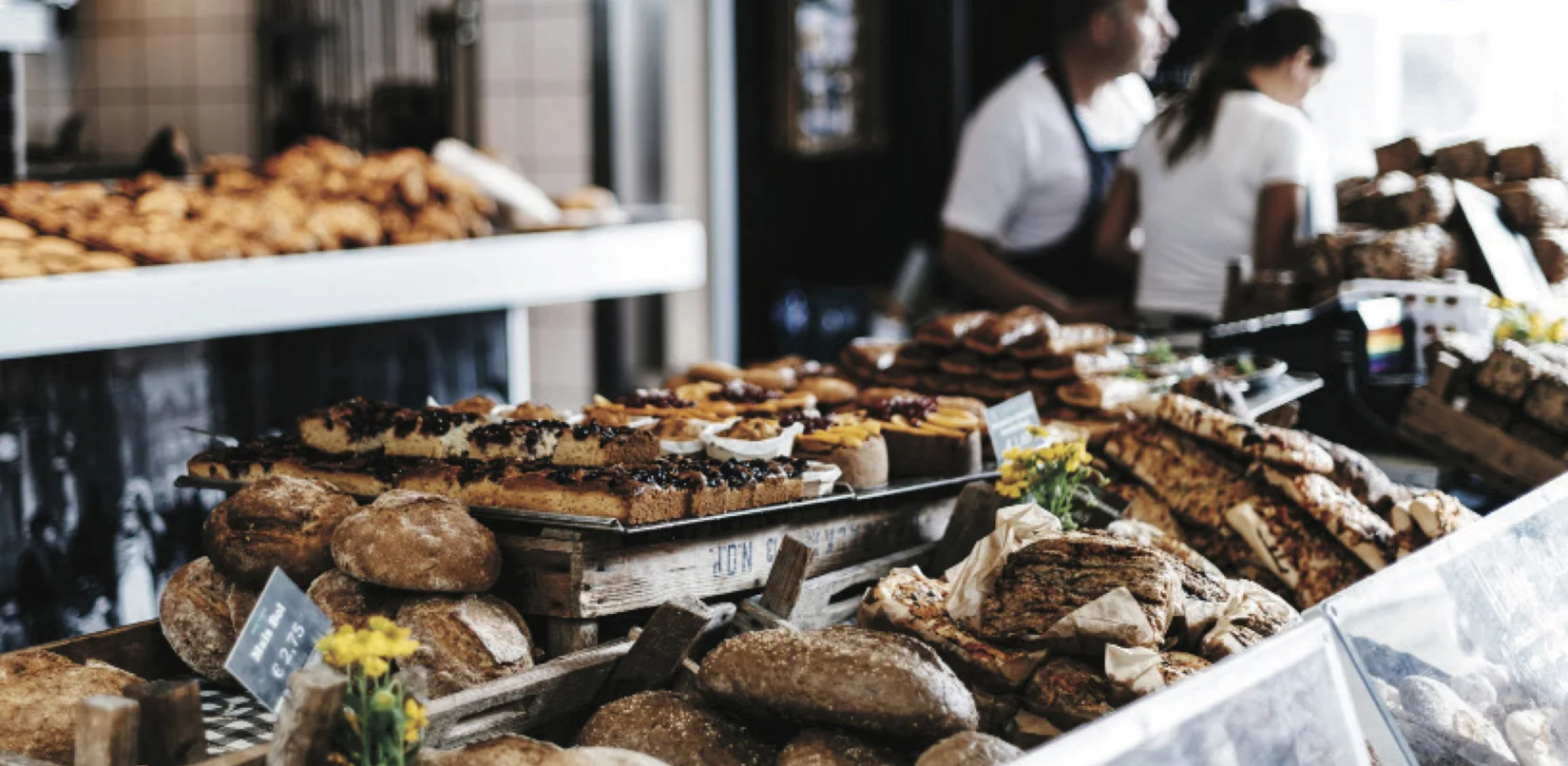 The busy Bake Culture display counter filled with breads, pastries and cakes, with two staff members behind it