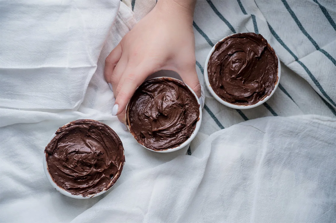 Overhead view of a hand holding a white ramekin of chocolate mousse beside two more ramekins on white linen