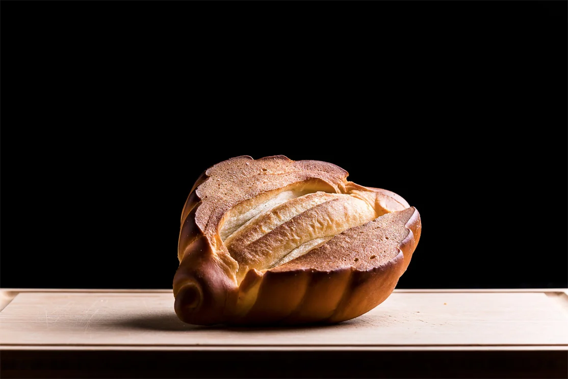 A single seashell-shaped bread loaf resting on a light wooden plank against a solid black background