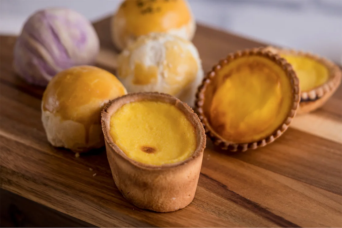 Egg tarts and assorted Asian pastry buns arranged on a wooden board