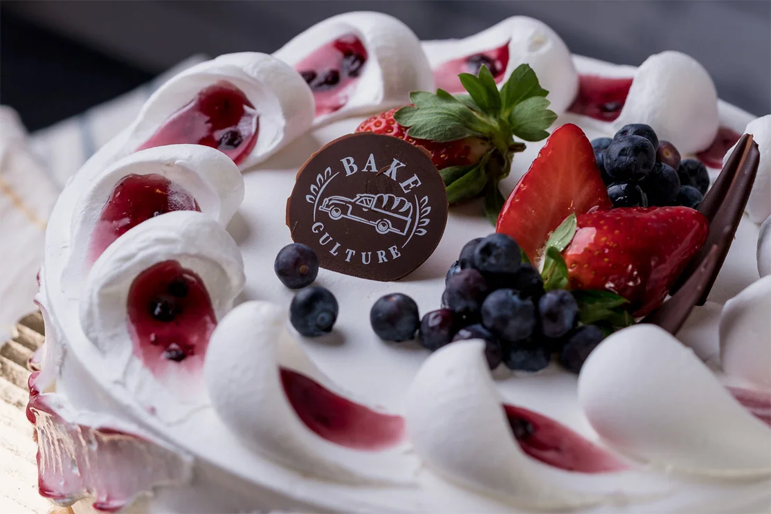 White cream cake with berry filling, meringue rosettes, strawberries and blueberries topped with a Bake Culture chocolate disc