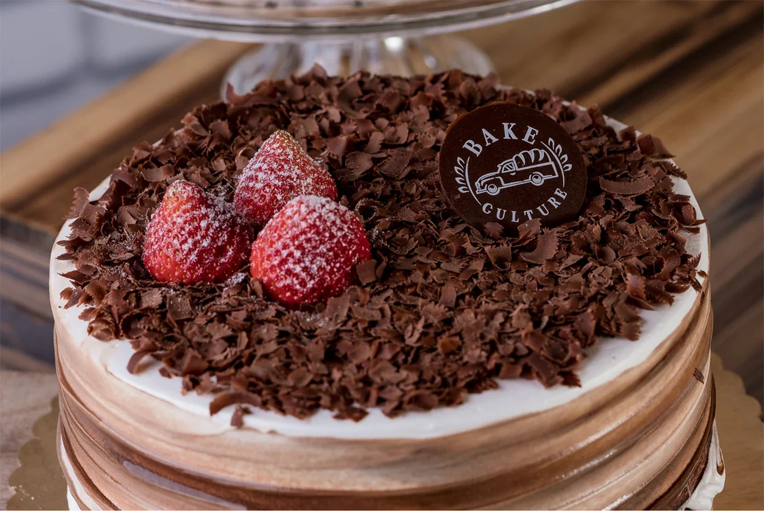 Chocolate layer cake with chocolate shavings, strawberries and a Bake Culture chocolate disc on a glass cake stand
