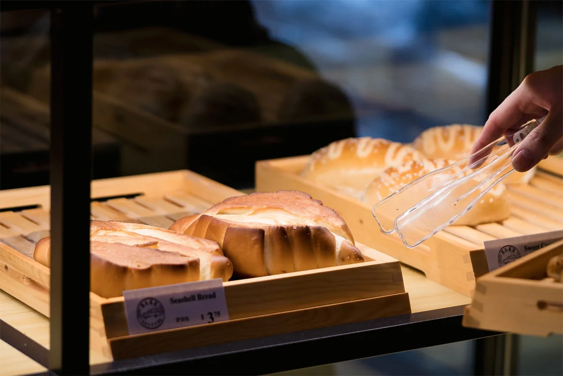 Close-up of the Bake Culture display case with seashell bread on wooden trays and a hand reaching in with tongs