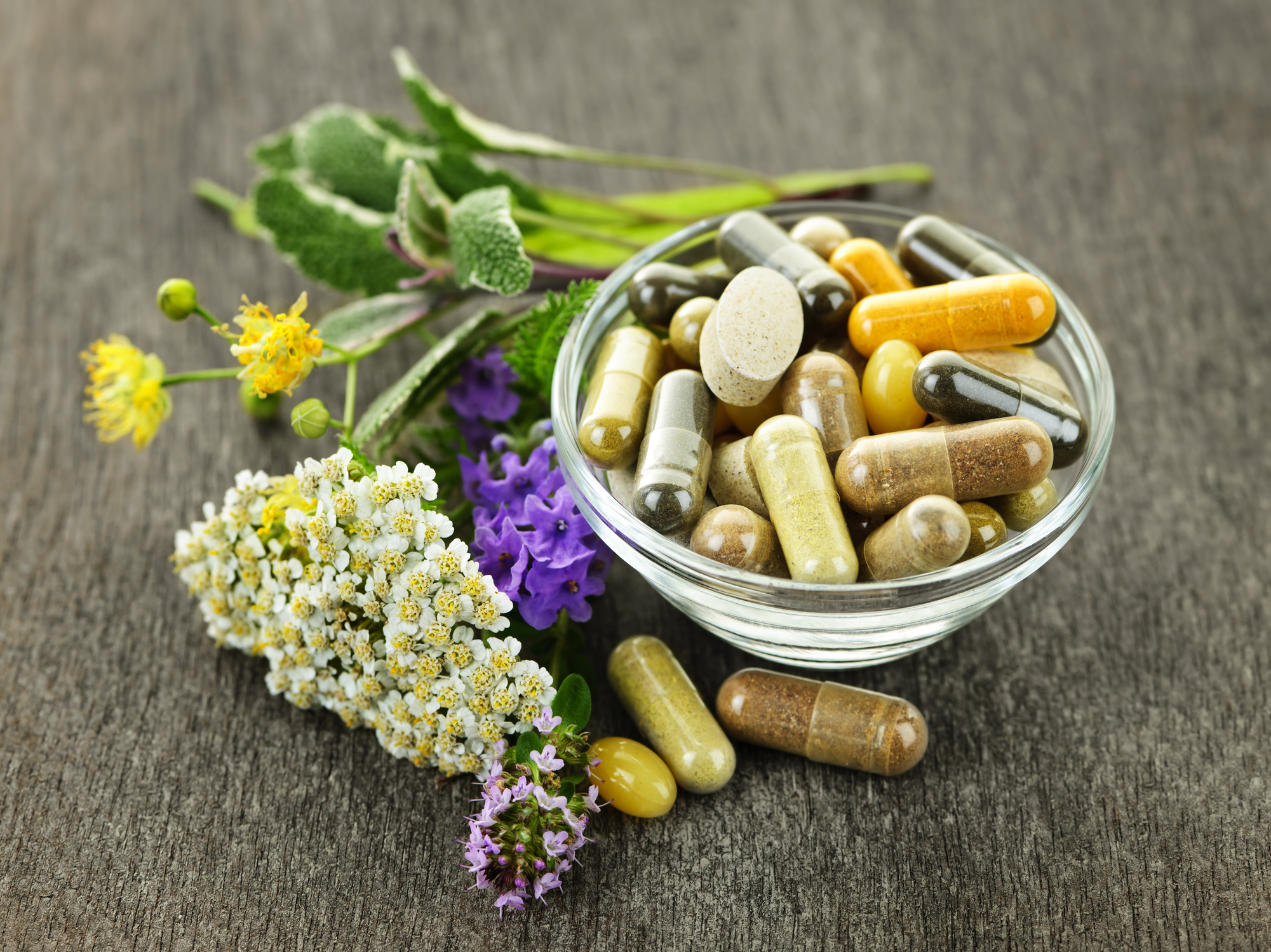A glass bowl of supplement capsules surrounded by herbs and wildflowers