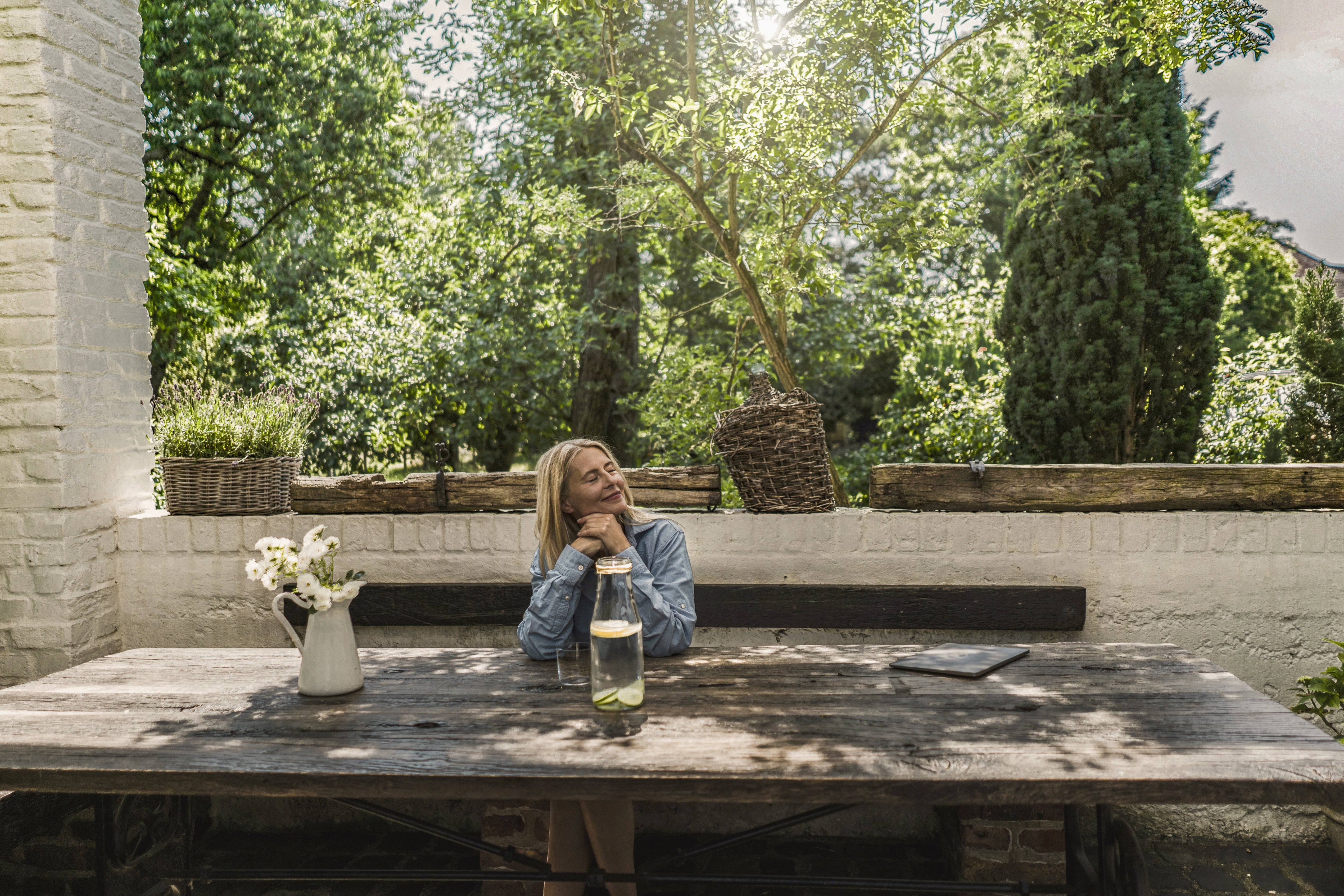 A woman in a denim shirt resting her chin on her hands at an outdoor wooden table with a jar of infused water