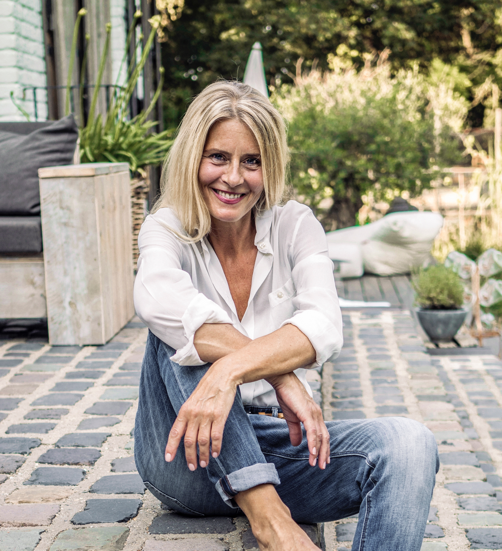 A smiling blonde woman in a white blouse and jeans sitting on a cobblestone patio surrounded by greenery