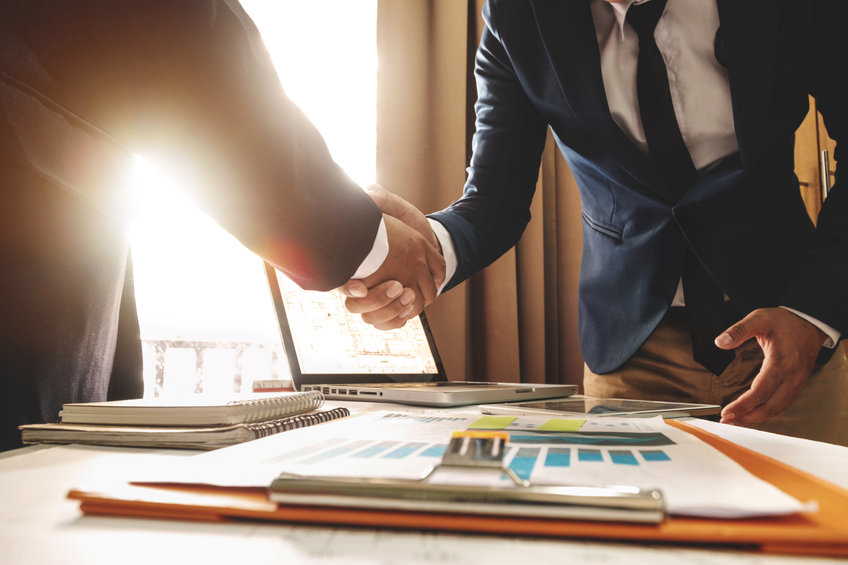 Two businessmen shaking hands over a desk with charts and a laptop