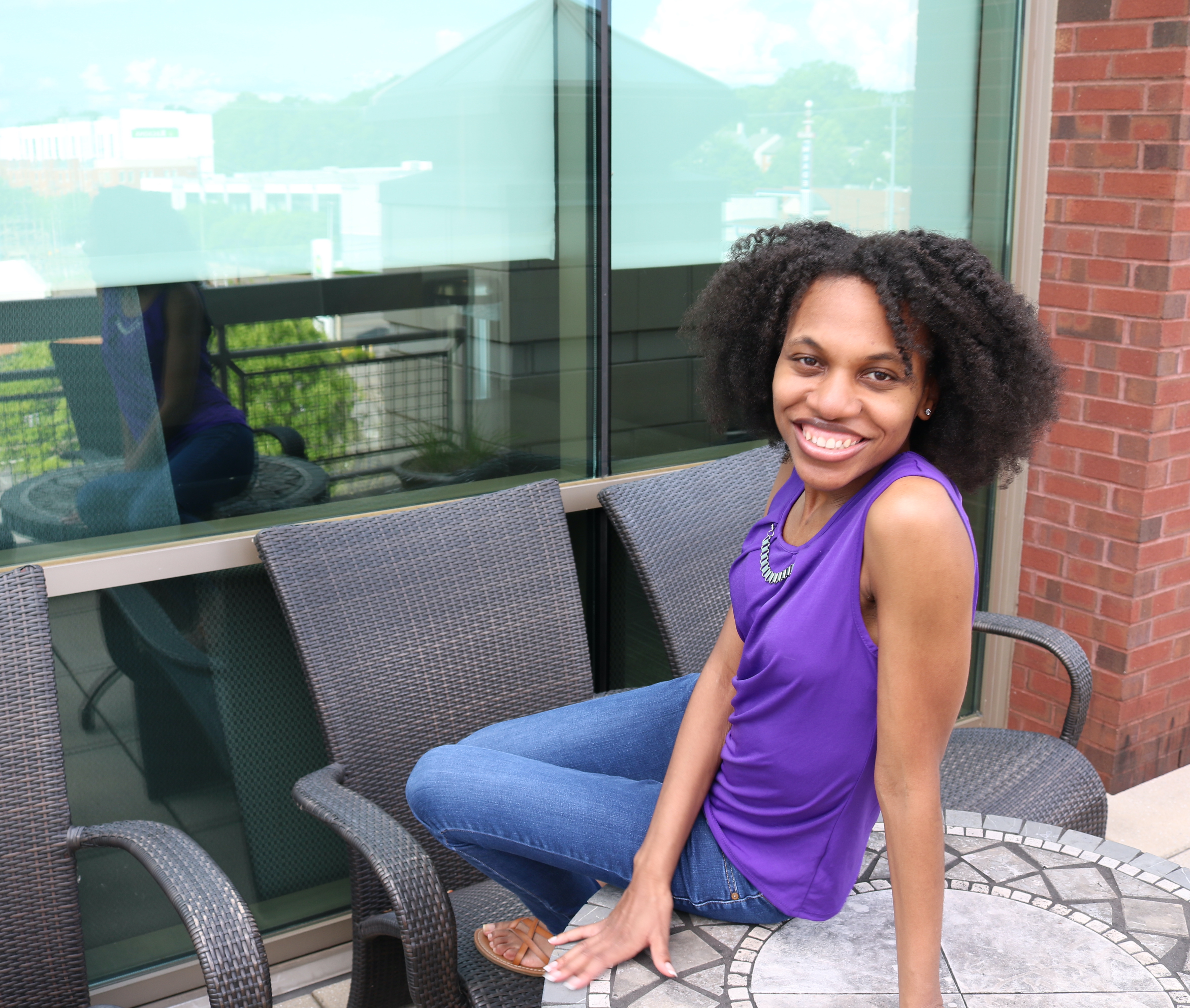 Dr. Lisa Battle-Gwathney seated on outdoor patio furniture in a purple tank top, smiling, with a glass building behind her.