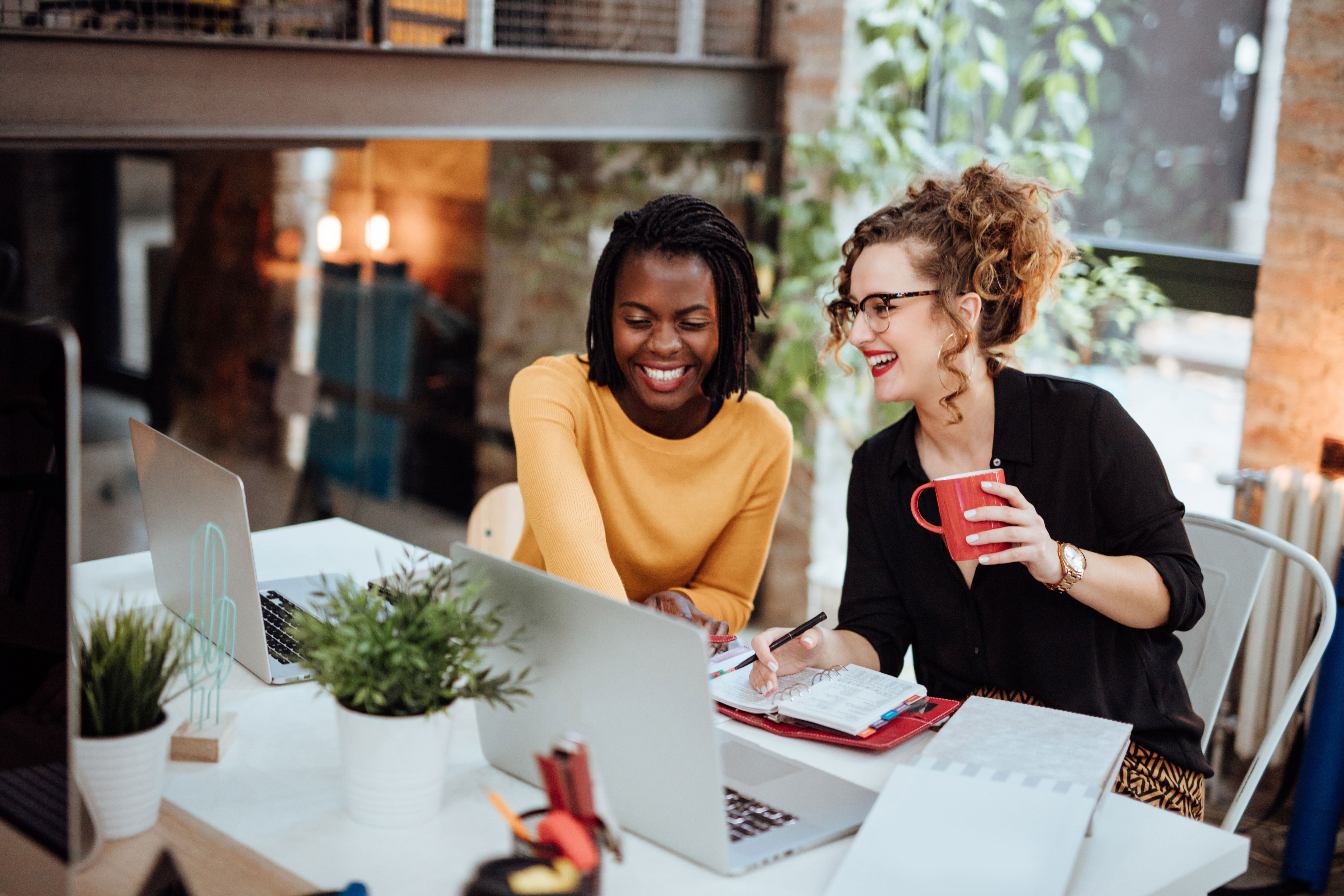 Two women laughing together at a desk with laptops and notebooks.