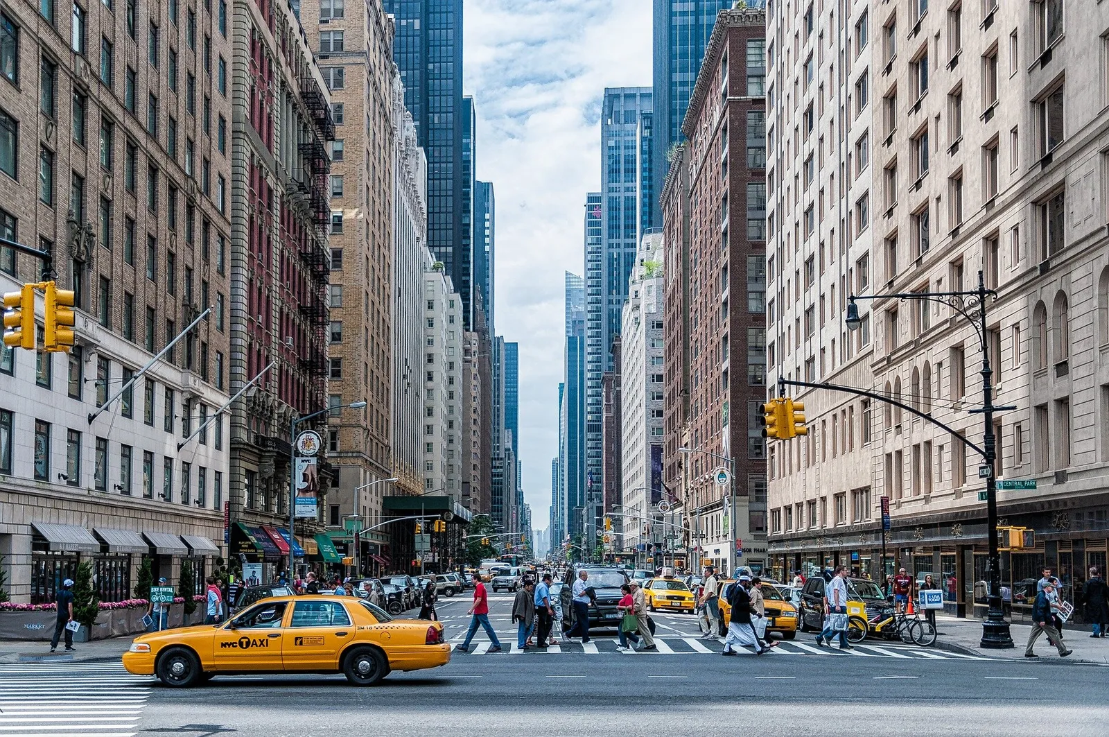 Busy New York City street with yellow taxi cabs, pedestrians crossing, and tall buildings lining both sides