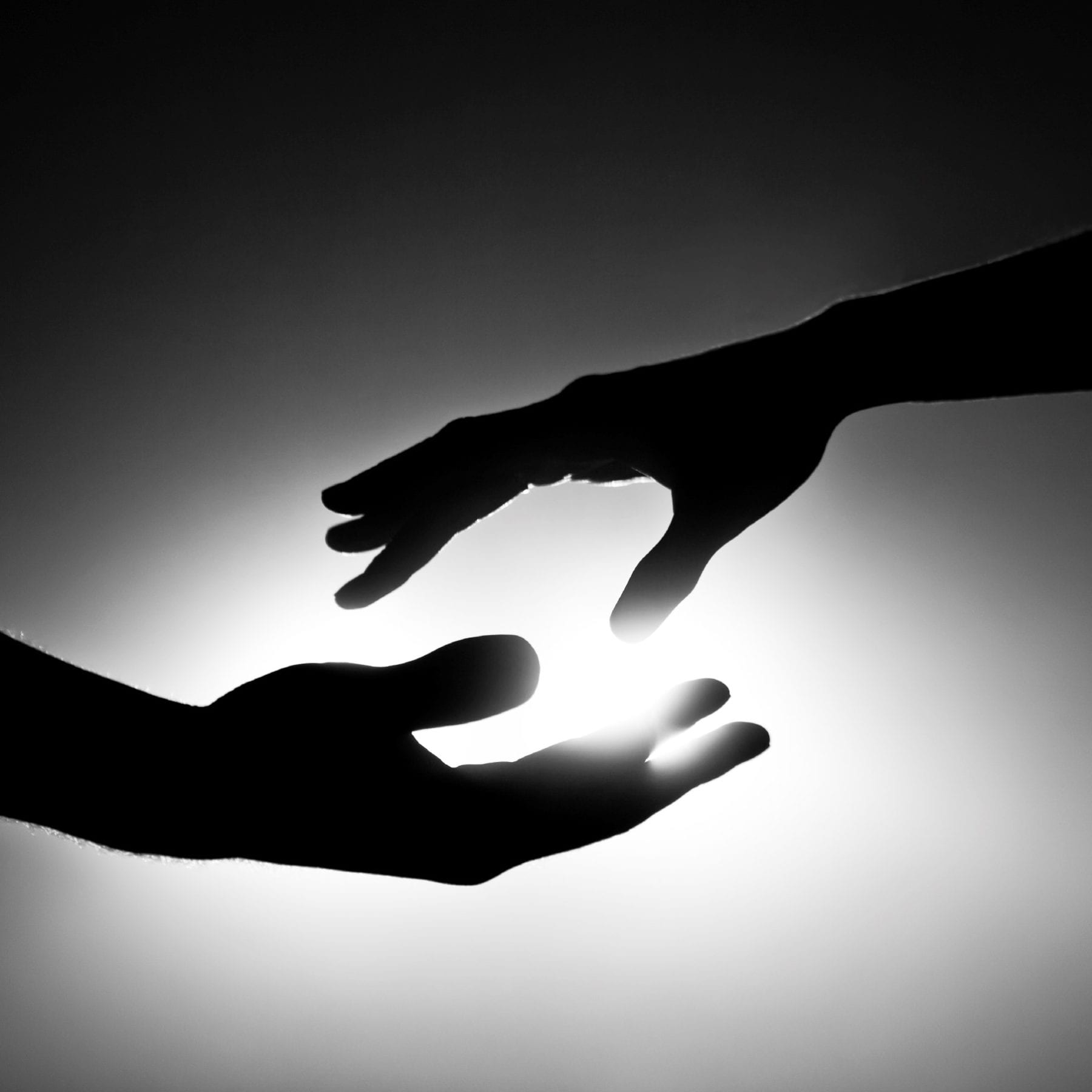 Black-and-white silhouette of two hands reaching toward each other against a bright light background