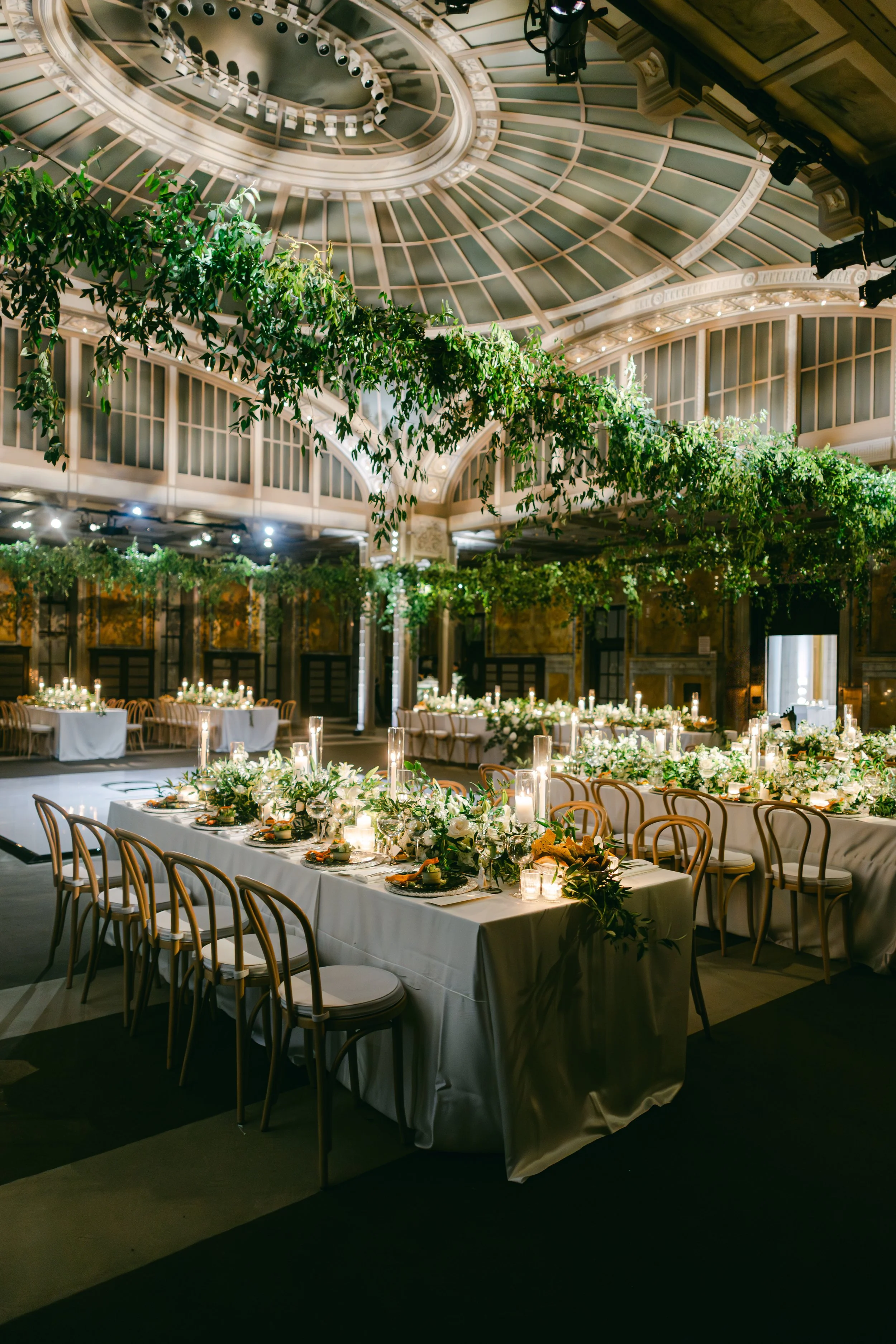 Elegant glass-domed reception hall with hanging greenery and long candlelit tables.