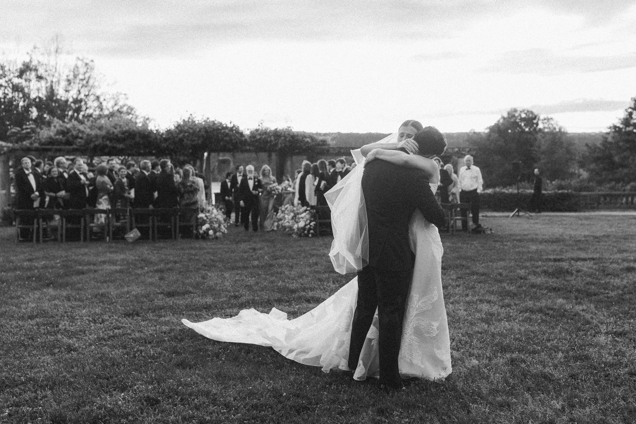 Groom lifting his bride after their outdoor ceremony, guests seated on the lawn behind them.