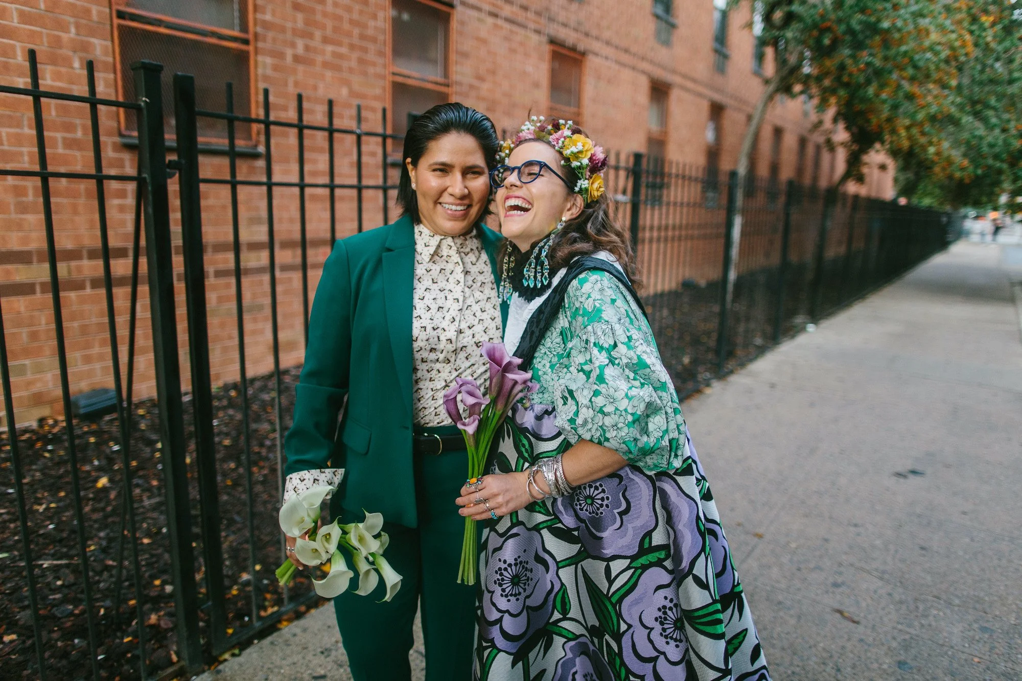 Two brides laughing on a New York City sidewalk, one in a green suit and one in a floral dress, holding calla lilies.