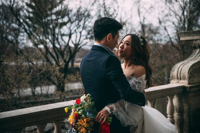 Couple embracing on a balcony with a colorful bouquet, bride in a lace off-shoulder gown.
