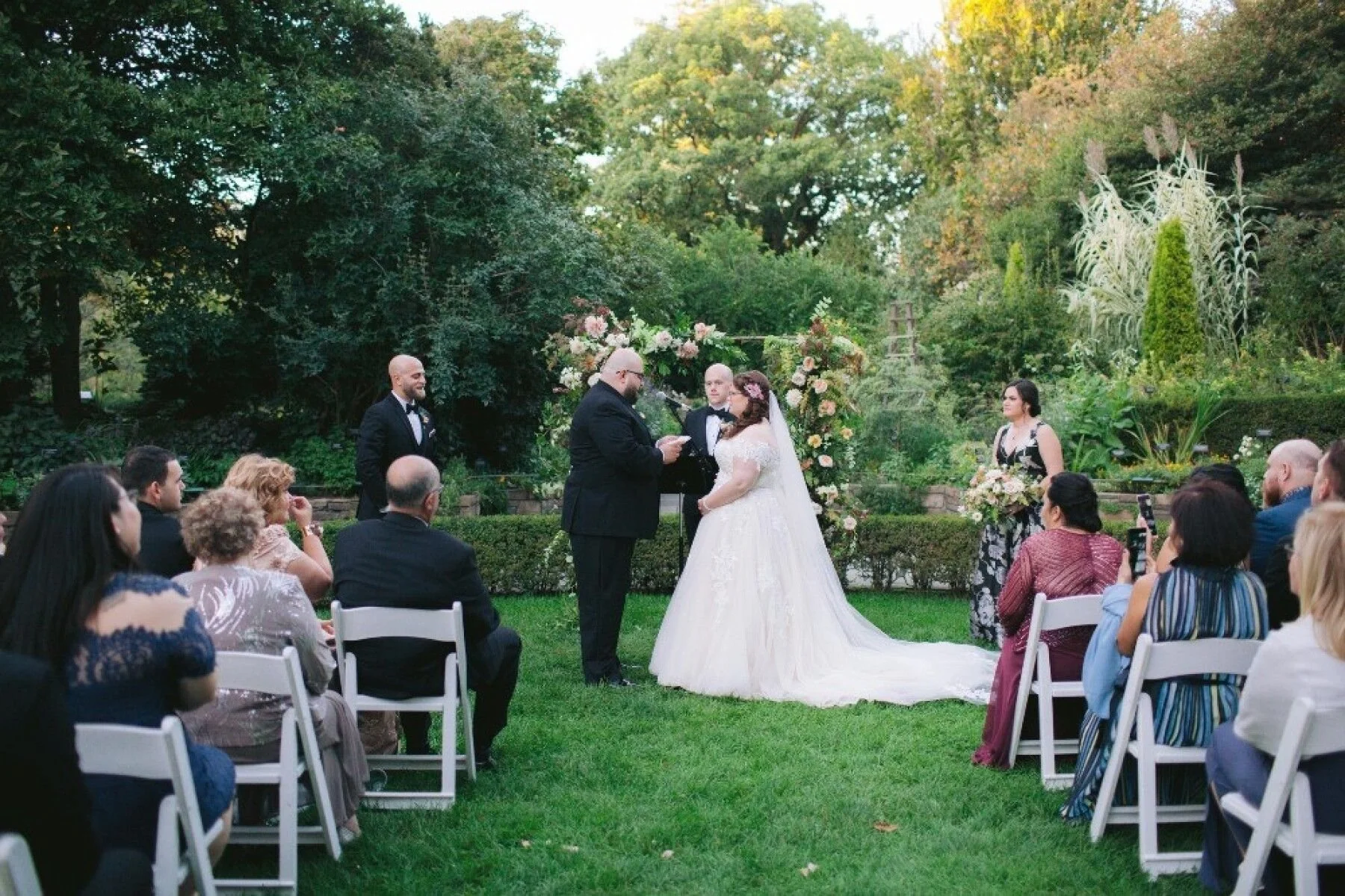 Outdoor garden ceremony, couple exchanging vows under a floral arch with seated guests.