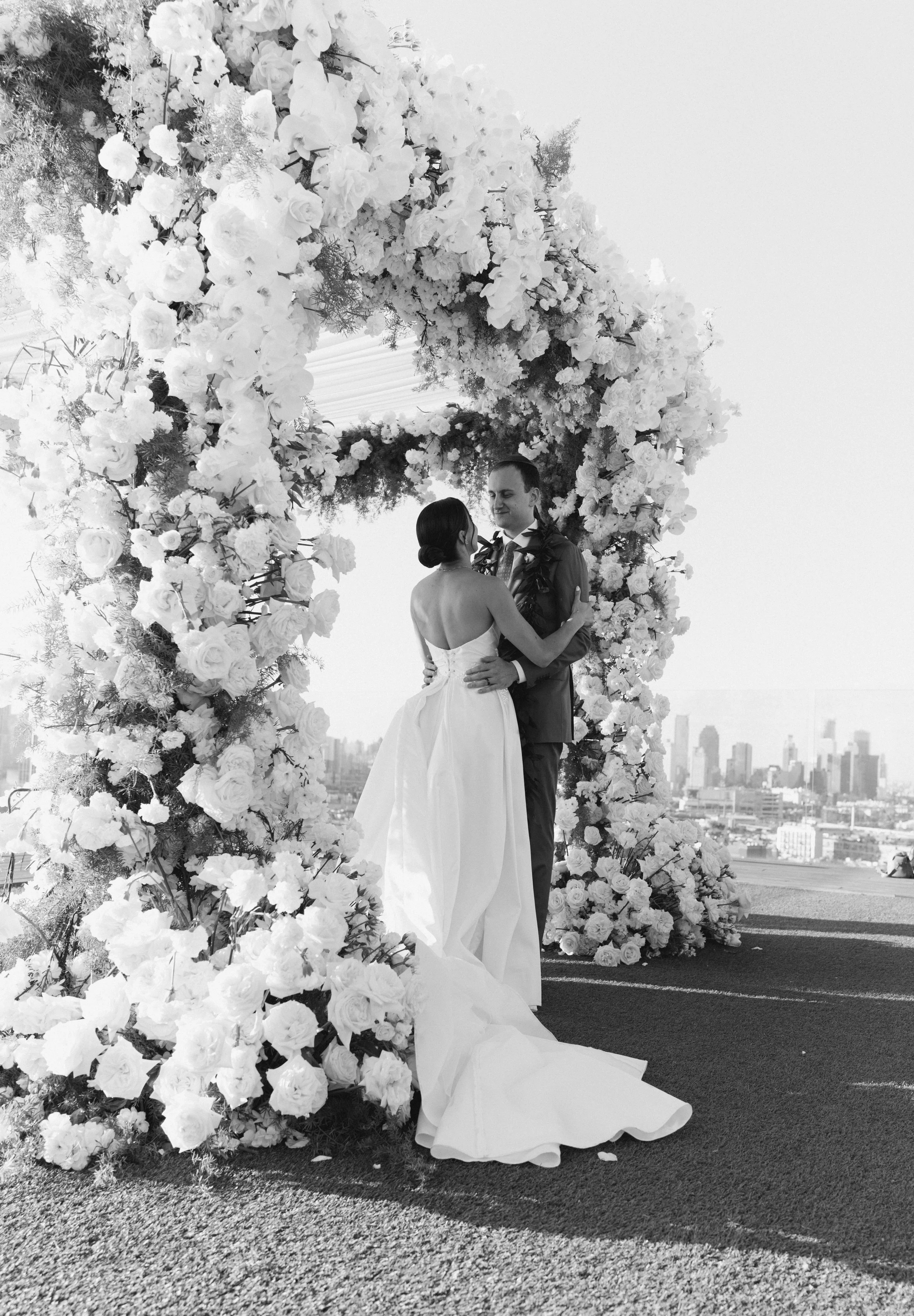 Black and white photograph of a bride and groom embracing under a white floral arch with the city skyline behind them.
