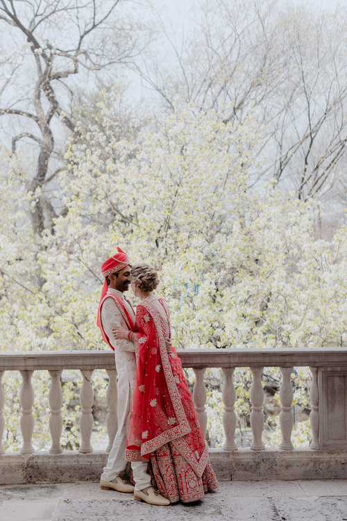 Indian wedding couple, groom in ivory sherwani and red turban with bride in a red lehenga, among spring blossoms.