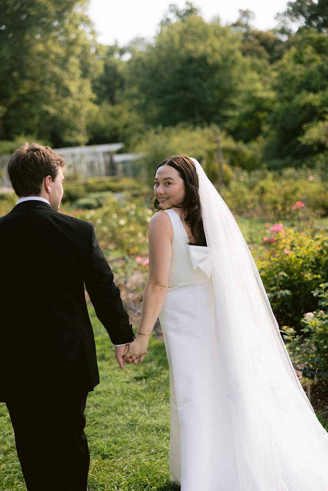 Bride in a veil looking back at her groom while walking through a garden.