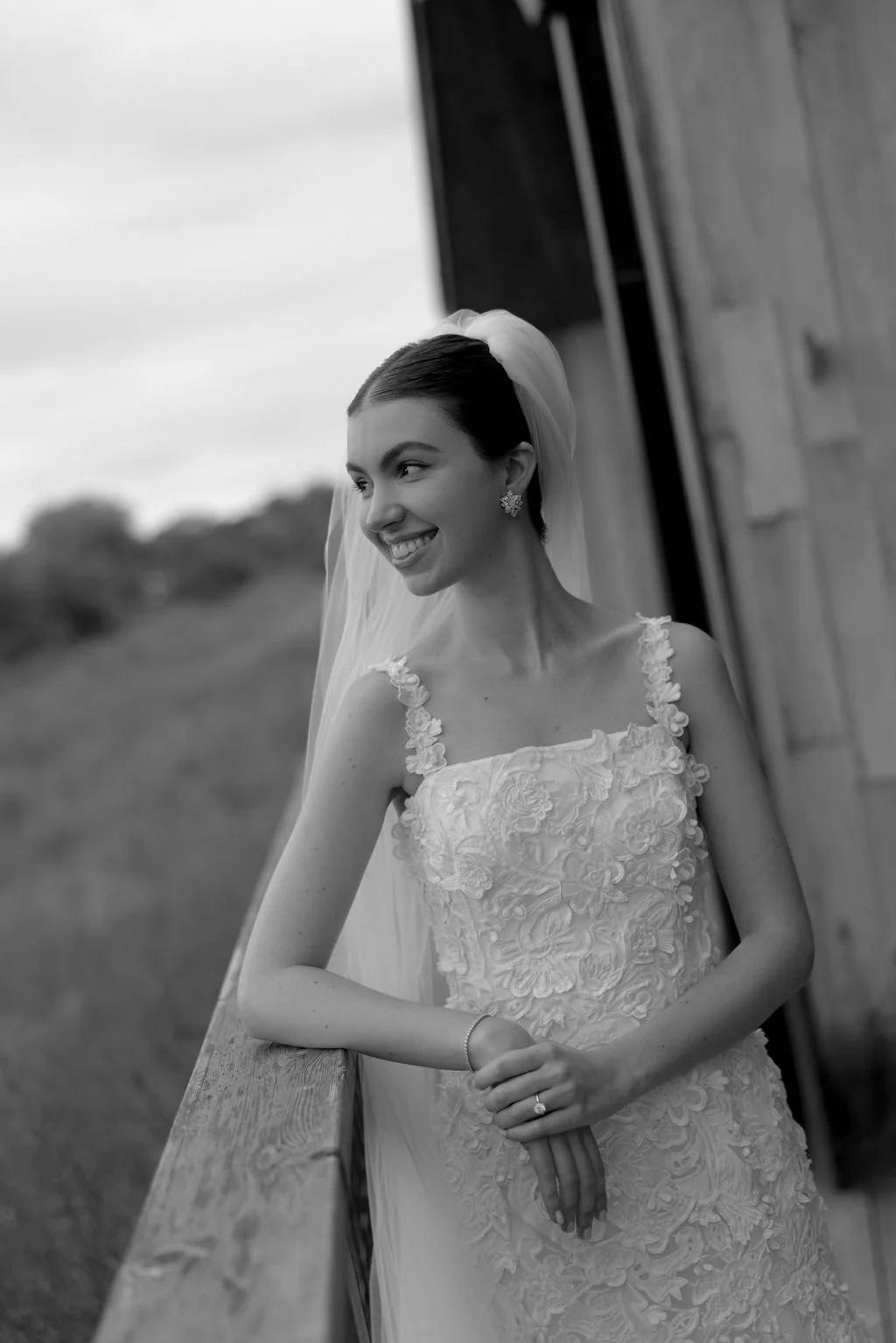 Black and white portrait of a bride in lace and veil leaning against a barn wall.