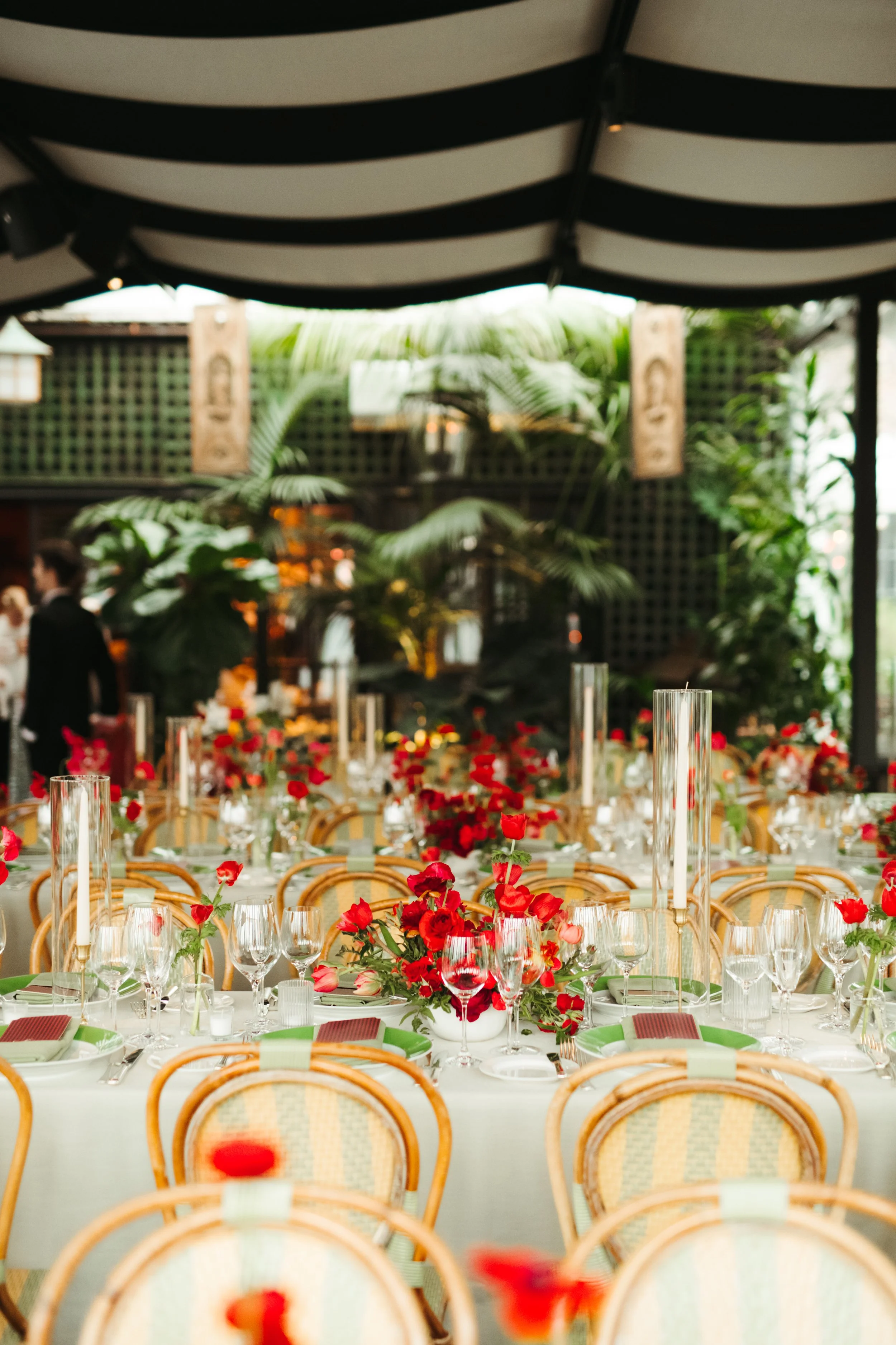 Outdoor reception under a striped awning with red floral centerpieces on round tables.