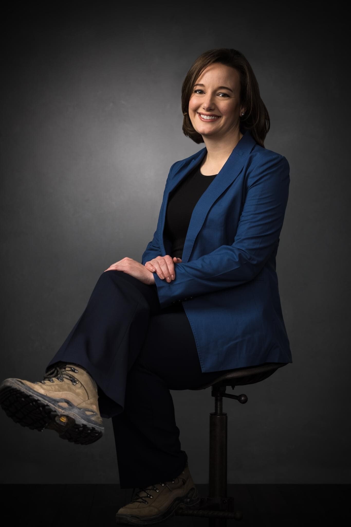 Laurie Hazel Patton in a blue blazer and hiking boots, seated on a stool against a dark studio background