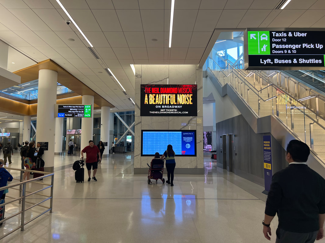 LGA airport interior with digital screens showing 'A Beautiful Noise' Broadway musical ad on a column