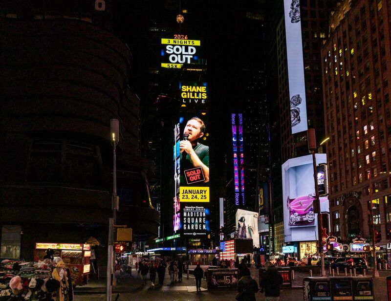 Shane Gillis 'Sold Out' Times Square digital billboard at night, wide shot of the square