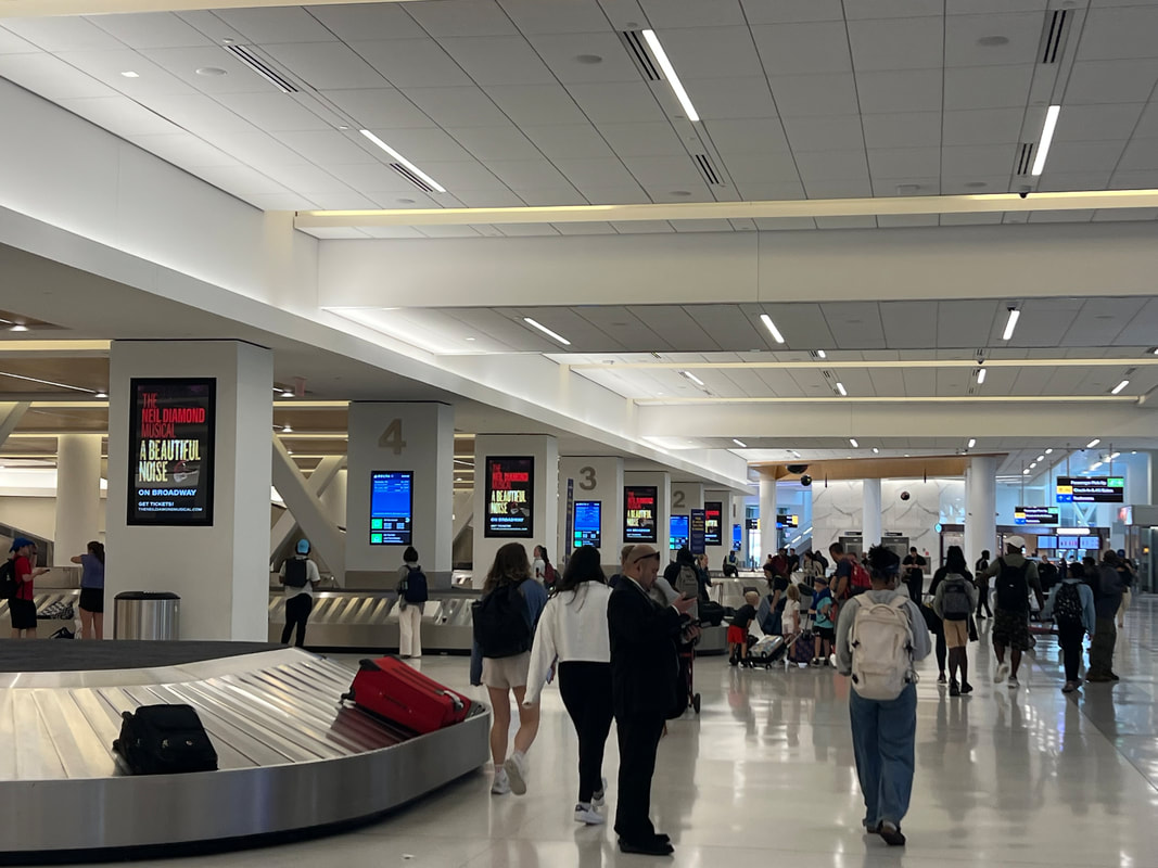 LGA airport baggage claim with multiple 'A Beautiful Noise' digital screens on columns