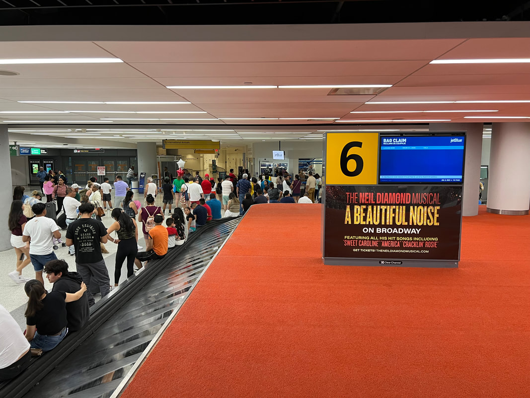 JFK airport baggage claim with 'A Beautiful Noise' diorama display kiosk beside carousel