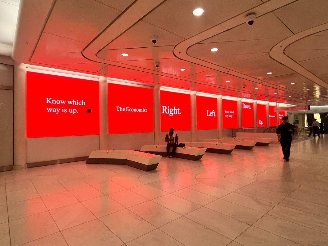 Row of The Economist red digital screens lining WTC corridor with benches and pedestrians
